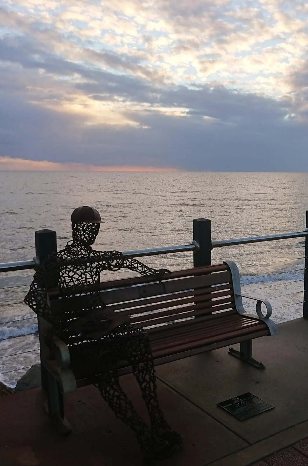 Timber-slatted public bench with a wire sculpture on a coastal foreshore at dusk